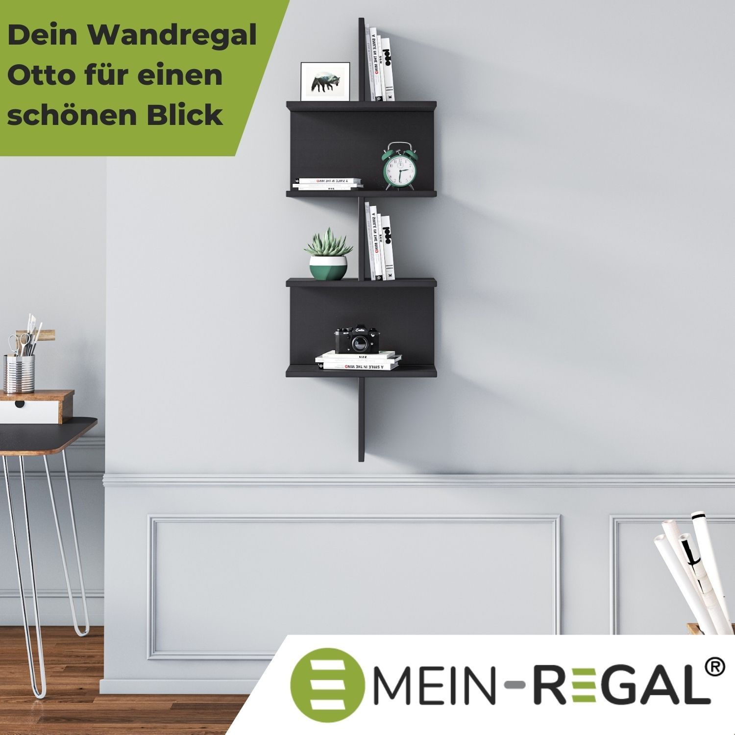 Wandregal Otto Schwarz im Büro neben einem Schreibtisch an grauer Wand hängend. Im Regal stehen Bücher, eine Kaktuspflanze, Fotoapparat, Wecker.